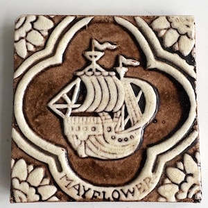May include: Square ceramic tile with a brown background and a white, stylized image of a sailing ship. The word "MAYFLOWER" is written in white at the bottom. The tile is framed with a white floral design.