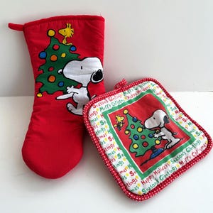 May include: A red Christmas stocking and a matching pot holder featuring Snoopy and Woodstock decorating a Christmas tree. The stocking and pot holder have a festive design with the words "Seasons Greetings" printed around the edges.