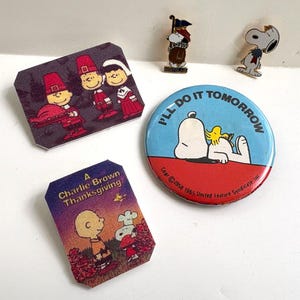 May include: A collection of vintage Peanuts memorabilia, including pins and magnets. The items feature Snoopy, Charlie Brown, and other characters in Thanksgiving and everyday scenes. One button reads "I'll do it tomorrow."