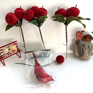 Peut inclure: Une collection d'objets décoratifs comprenant trois tiges de pommes rouges artificielles avec des feuilles vertes, un petit banc en métal rouge avec deux oursons miniatures, un oiseau rouge dans un sachet plastique et un ours en peluche marron portant un bonnet et tenant un panneau indiquant BLESS THIS HARVEST.