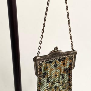 Peut inclure: Sac &agrave; main ancien en m&eacute;tal maill&eacute; avec un cadre d&eacute;coratif et une cha&icirc;ne. Le sac pr&eacute;sente un motif de carr&eacute;s noirs, jaunes et verts. Le bord inf&eacute;rieur a une frange de brins m&eacute;talliques. Le sac est expos&eacute; sur un support noir.