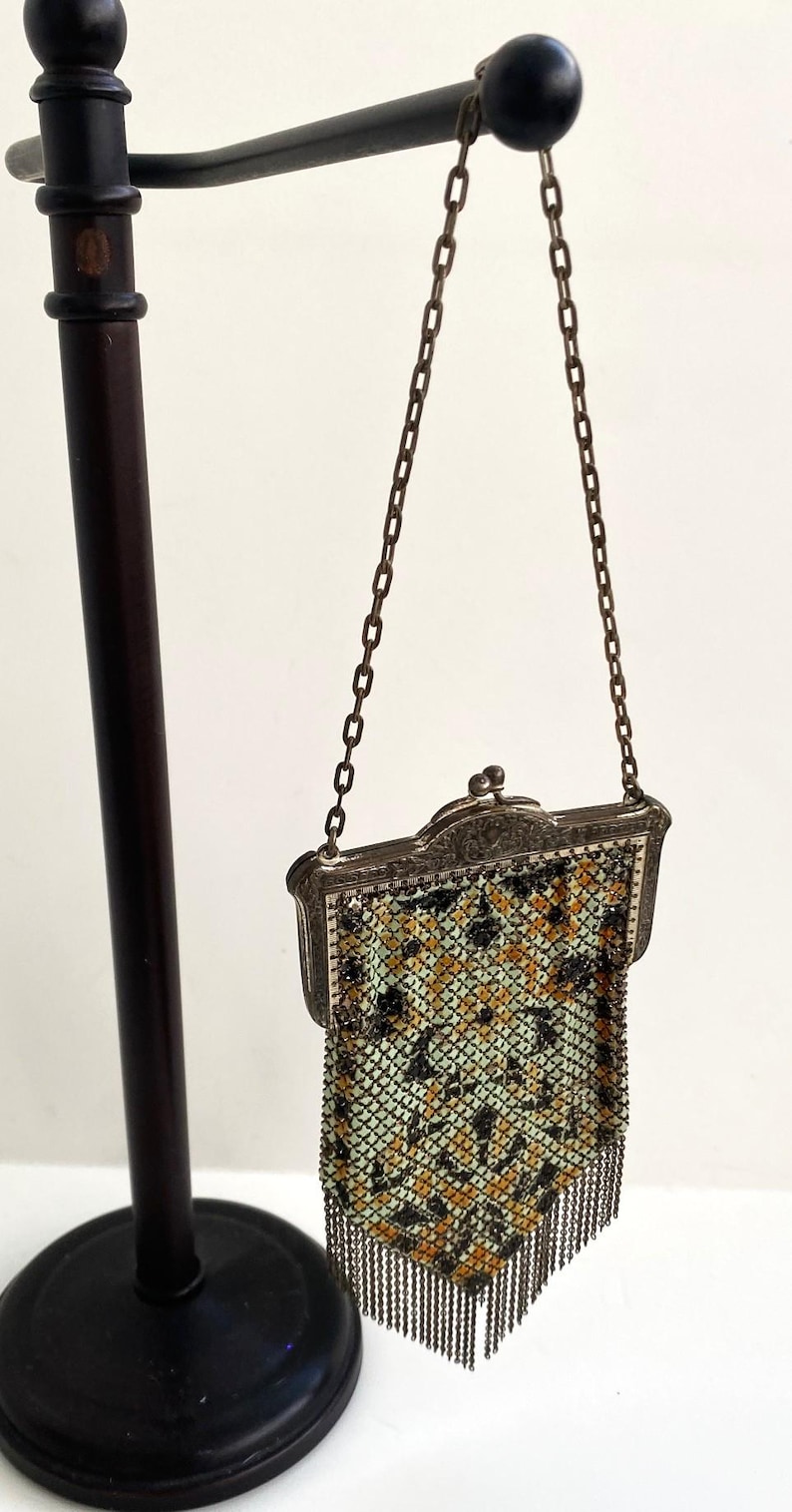 Peut inclure: Sac &agrave; main ancien en m&eacute;tal maill&eacute; avec un cadre d&eacute;coratif et une cha&icirc;ne. Le sac pr&eacute;sente un motif de carr&eacute;s noirs, jaunes et verts. Le bord inf&eacute;rieur a une frange de brins m&eacute;talliques. Le sac est expos&eacute; sur un support noir.