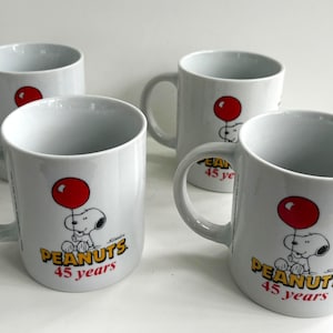 May include: Four white ceramic mugs featuring Snoopy holding a red balloon. Each mug has the text "PEANUTS 45 years" in gold and red. The mugs have a curved handle and a glossy finish.