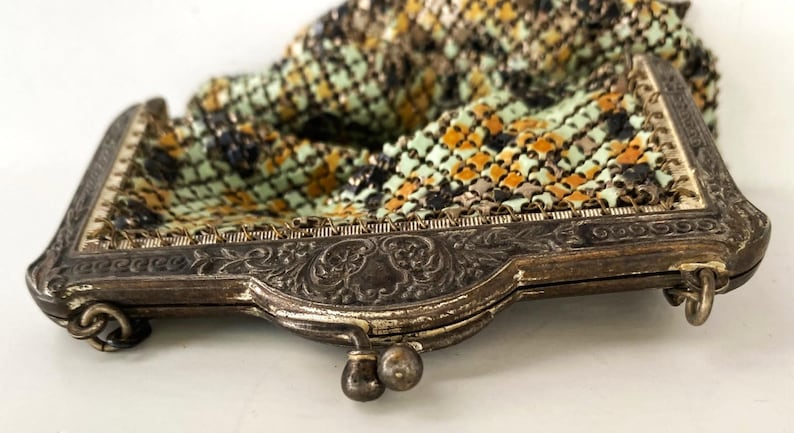 Peut inclure: Un sac &agrave; main ancien en m&eacute;tal maill&eacute; avec un cadre d&eacute;coratif argent&eacute;. Le sac pr&eacute;sente un motif color&eacute; de vert, orange et noir. Le cadre a des d&eacute;tails floraux orn&eacute;s et une fermeture &agrave; fermoir.