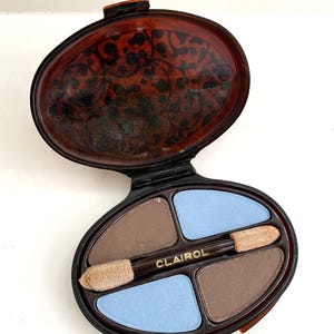 May include: A vintage oval-shaped compact with a brown tortoiseshell design. The compact opens to reveal four eyeshadows in shades of brown and blue. The compact also includes two applicators and the word "CLAIROL" printed in gold.