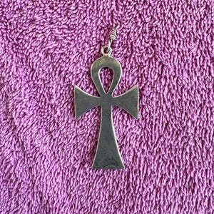 Peut inclure: Un pendentif ankh en métal argenté présentant une surface lisse et plate avec un sommet en boucle et une base évasée. Le pendentif comprend un petit anneau de jonction en métal au sommet, fixé à une boucle en fil d'argent torsadé.