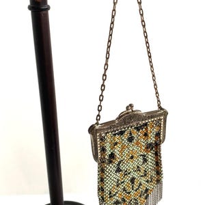 Peut inclure: Un sac &agrave; main ancien en m&eacute;tal maill&eacute; avec un cadre d&eacute;coratif et une cha&icirc;ne. Le sac pr&eacute;sente un motif de carr&eacute;s noirs, jaunes et verts. Il est pr&eacute;sent&eacute; sur un support marron fonc&eacute;.