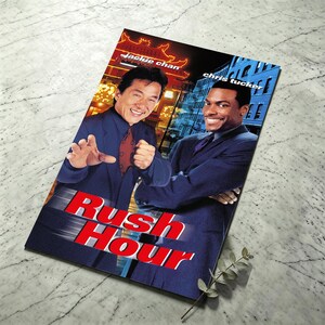 Pode incluir: Um cartaz de filme retangular de Rush Hour com Jackie Chan e Chris Tucker em fatos azul-escuro. O título Rush Hour está impresso em letras vermelhas grandes na parte inferior. O fundo mostra uma cena de rua com iluminação quente.