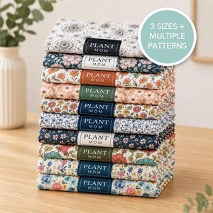 May include: A stack of ten folded fabric items with various floral patterns, each featuring a rectangular label that reads PLANT MOM. A circular graphic in the upper right corner displays the text 3 SIZES + MULTIPLE PATTERNS in white.