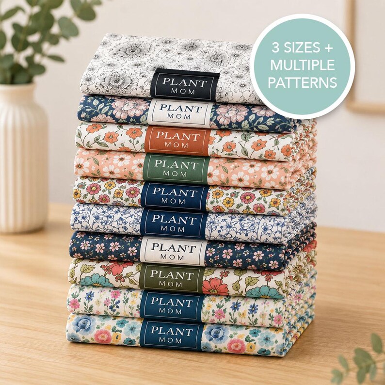 May include: A stack of ten folded fabric items with various floral patterns, each featuring a rectangular label that reads PLANT MOM. A circular graphic in the upper right corner displays the text 3 SIZES + MULTIPLE PATTERNS in white.