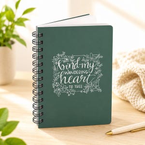 May include: A dark green spiral-bound notebook featuring white line art of flowers and leaves surrounding the hand-lettered text "bind my wandering heart TO THEE" centered on the front cover. The notebook has a black metal coil binding.