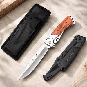 May include: A folding knife with a silver blade featuring three circular cutouts and a wood-grain handle engraved with the name James. Beside it lies a black nylon sheath and a closed black folding knife with the name Austin printed on the side.