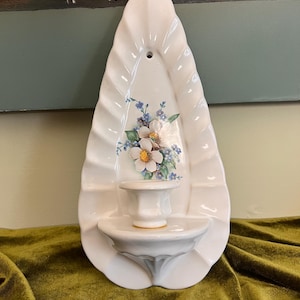 May include: A white ceramic wall sconce featuring a teardrop-shaped backplate with a fluted edge and a central floral decal of white blossoms and blue forget-me-nots. The piece includes a small integrated shelf and a single candle holder cup.
