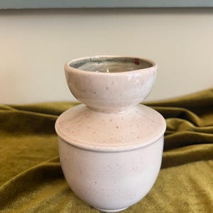 May include: A ceramic butter dish with a lid, featuring a light cream-colored glaze with subtle brown speckles throughout. The piece has a rounded base and a matching lid with a wide, circular handle at the top, creating a smooth, uniform appearance.