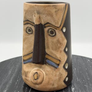 May include: A cylindrical ceramic mug featuring a stylized face with a prominent dark brown nose, circular eyes, and a small mouth. The surface has a tan, textured finish with dark brown accents, blue horizontal lines, and a side panel with white dots.