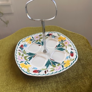 May include: A white ceramic serving tray with four circular indentations and a central silver metal handle. The tray features a colorful floral pattern with yellow, pink, and red flowers, green leaves, and a scalloped green edge along the border.