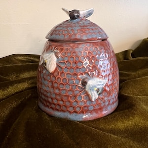 May include: A ceramic honey pot with a lid featuring a sculpted bee handle. The pot has a textured honeycomb pattern in shades of reddish-brown and blue, with two additional sculpted bees attached to the exterior surface of the container.