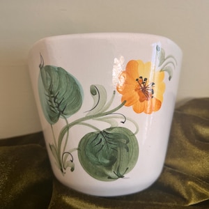 May include: A white ceramic planter featuring a hand-painted design of a single orange flower with dark brown centers and two large green leaves with detailed brushstrokes. The pot has a smooth, glossy finish and a tapered, cylindrical shape.