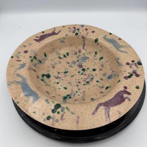 May include: A round ceramic bowl with a speckled beige base featuring scattered purple and green paint splatters. The wide rim is decorated with silhouettes of horses in shades of purple and light blue, arranged around the circular edge.
