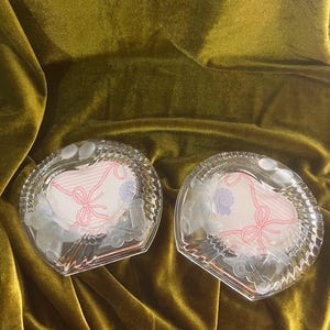 May include: Two clear glass dishes with a scalloped edge and a central circular inset. The inset features a white background with a pink line-drawn bow and ribbon design. The glass border is decorated with molded figures of a teddy bear and a train.