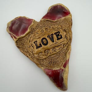 May include: A heart-shaped ceramic wall plaque featuring a tan, textured surface with intricate lace-like patterns and the word LOVE embossed in the center. The edges and top sections are finished with a glossy, deep red glaze.