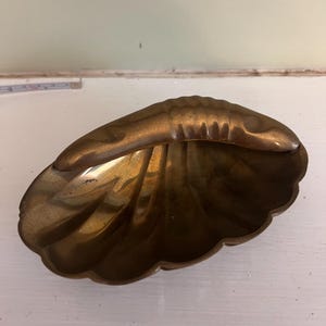 May include: A brass dish shaped like a scallop shell with a raised lobster figure attached to the top edge. The dish has a smooth, metallic finish with visible ridges radiating from the center. A partial measuring tape is visible in the background.