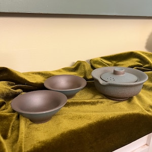 May include: A ceramic tea set consisting of two small, wide-rimmed bowls and a lidded teapot with a side spout and a built-in strainer. The pieces feature a matte, textured finish with a muted blue-green exterior and a dark brown interior.