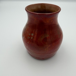 May include: A small, bulbous ceramic vase with a glossy, deep red glaze. The surface features subtle variations in color, including darker reddish-brown tones and faint, lighter circular markings. The vase has a wide, open mouth and a smooth, rounded body.
