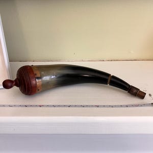 Può includere: Un corno da polvere curvo con un corpo in corno scuro lucidato, una base in legno arrotondata con pomello e una punta in metallo. Un metro a nastro bianco è posizionato sotto, indicando una lunghezza totale di circa 33 cm.