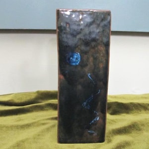 May include: A rectangular ceramic vase with a dark, mottled glaze featuring shades of black, brown, and deep blue. The surface has a glossy finish with a prominent circular blue accent near the top and a thin, vertical blue line extending downward.