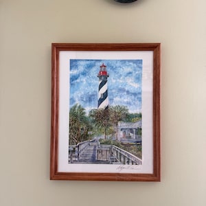 May include: A framed watercolor painting depicting a black and white spirally striped lighthouse under a blue sky. A wooden boardwalk leads toward the lighthouse, flanked by green palm trees and a small building. The artist's signature is at the bottom.