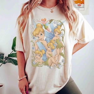 Könnte beinhalten: Ein cremefarbenes Kurzarm-T-Shirt mit einem Grafikdruck von vier Tinker Bell-Figuren in verschiedenen Posen. Das Design enthält grüne und blaue Akzente mit wirbelnden, rankenartigen Mustern, die die Figuren auf der Vorderseite des Shirts umgeben.