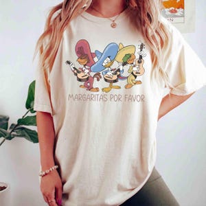 Pode incluir: Uma t-shirt de manga curta na cor creme com um gráfico de três pássaros de desenho animado usando sombreros e tocando violão. Abaixo da ilustração, o texto MARGARITAS POR FAVOR está impresso em uma fonte simples de cor marrom.