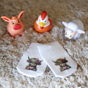 May include: A pair of white baby socks featuring a brown and white highland cow design. The socks are displayed on a fluffy white rug with toy farm animals.