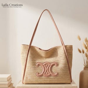 K&ouml;nnte beinhalten: Eine geflochtene Strohtasche mit hellbraunem, strukturiertem K&ouml;rper und glatten, rosafarbenen Lederriemen. Auf der Vorderseite befindet sich ein auff&auml;lliges, rosafarbenes Lederemblem mit einem symmetrischen, stilisierten Design und offener Form.