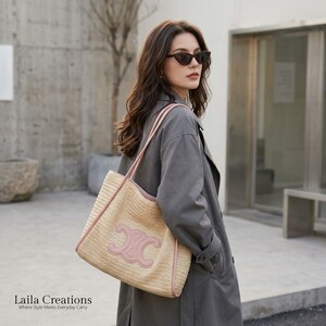 K&ouml;nnte beinhalten: Eine geflochtene Strohtasche mit einem gro&szlig;en, hellrosa, symmetrischen Emblem auf der Vorderseite. Die Tasche verf&uuml;gt &uuml;ber zwei passende hellrosa Schulterriemen und eine hellrosa Einfassung an den Kanten. Die Strohstruktur ist deutlich erkennbar.