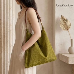 Pode incluir: Uma bolsa hobo grande verde-oliva com um padrão floral tonal subtil e uma alça de ombro de couro castanho com ferragens de metal dourado. A bolsa é usada no ombro. O texto no canto superior direito diz Laila Creations Where Style Meets Everyday Carry.