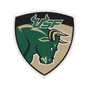 May include: A shield-shaped embroidered patch featuring a green bull head graphic on a tan background with a black upper section. The letters USF are stitched in green above the bull. The entire patch is outlined with a white border.