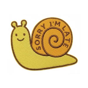 Könnte beinhalten: Ein gestickter Aufnäher in Form einer hellgelben Schnecke mit brauner Umrandung. Die Schnecke hat ein rundes orangefarbenes Gehäuse mit dem dunkelbraunen Text SORRY I'M LATE, der entlang der Spirale verläuft. Sie hat zwei kleine Fühler.