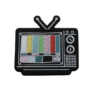 May include: An embroidered patch shaped like a vintage television set with a black frame and two top antennas. The screen displays a colorful vertical test pattern in yellow, blue, green, pink, and red, with a grid of smaller blocks below.
