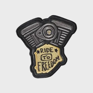 May include: An embroidered patch shaped like a motorcycle engine with a black border and grey engine details. A central tan shield-shaped area features the black embroidered text RIDE TO FREEDOM with a small star on each side of the word RIDE.