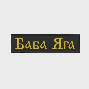 May include: A rectangular black embroidered patch featuring the text "БАБА ЯГА" stitched in yellow thread. The patch has a clean, straight-edged border and a flat, uniform surface, displaying the Cyrillic characters clearly against the dark background.