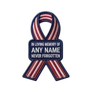 May include: A ribbon-shaped embroidered patch featuring a red, white, and blue striped pattern. A rectangular blue center section contains the white embroidered text: IN LOVING MEMORY OF ANY NAME NEVER FORGOTTEN. The patch has a dark blue border.