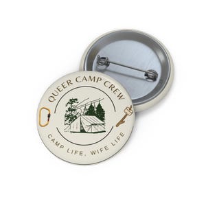 Könnte beinhalten: Ein runder Ansteckbutton mit cremefarbenem Hintergrund. Das Design zeigt ein Zelt, Bäume, einen Karabiner und einen Marshmallow am Stock. Der Text "QUEER CAMP CREW" und "CAMP LIFE, WIFE LIFE" ist kreisförmig um die Mitte angeordnet.