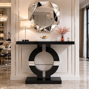 May include: A black console table with a circular base design and a rectangular top. Above the table hangs a large, multi-faceted geometric mirror. On the table surface sits a lamp with a white shade, a gold bowl, books, and a vase with pink flowers.