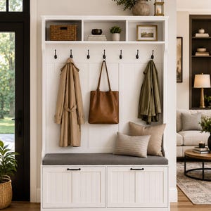 May include: A white hall tree with a bench, featuring a dark gray cushioned seat, two storage drawers with black handles, and six black coat hooks. The upper section includes three open storage cubbies holding a basket, decor, and a framed picture.