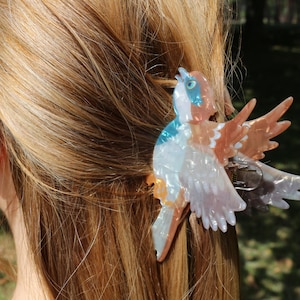 Puede incluir: Una pinza para el cabello con forma de pájaro con un acabado nacarado en tonos azul, blanco y marrón. El diseño del pájaro presenta alas extendidas, un pequeño detalle de ojo azul y un mecanismo de resorte metálico visible en el centro.