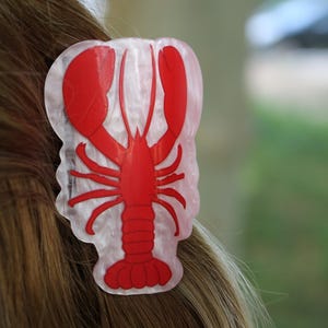 Puede incluir: Una pinza para el cabello con una silueta de langosta de color rojo brillante centrada sobre una base de acrílico translúcido de color blanco nacarado y veteado. El diseño de la langosta incluye dos grandes pinzas, patas y una cola segmentada.