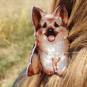 May include: A hair claw clip shaped like a German Shepherd dog. The accessory features a glossy, pearlescent finish with brown, tan, and white colouring. The dog design shows a smiling expression with upright ears, a black nose, and a pink tongue.
