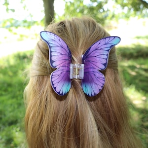 Puede incluir: Una pinza para el cabello con forma de mariposa que presenta un diseño degradado que va del morado oscuro en el centro al azul claro en los bordes de las alas. Las alas tienen un patrón detallado de venas negras y un mecanismo de bisagra transparente.